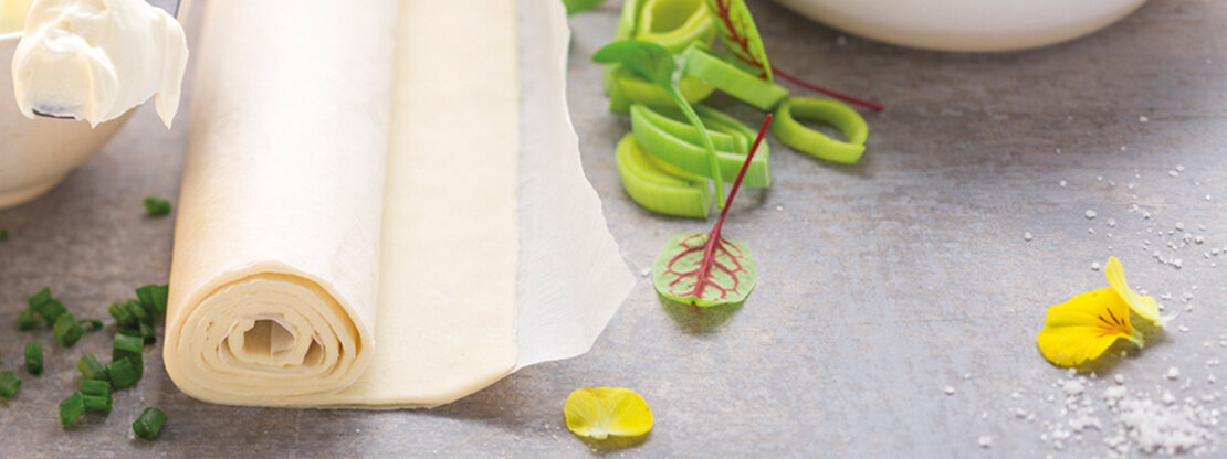 Picture of a Wewalka fresh flatbread dough, which is pre-rolled on baking paper, lying with pieces of leek on the worktop of a kitchen 