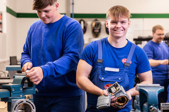 Two young men work in their apprenticeships at the work table. They are holding a motor and various small parts are spread around them. In the back their trainers are talking to each other.
